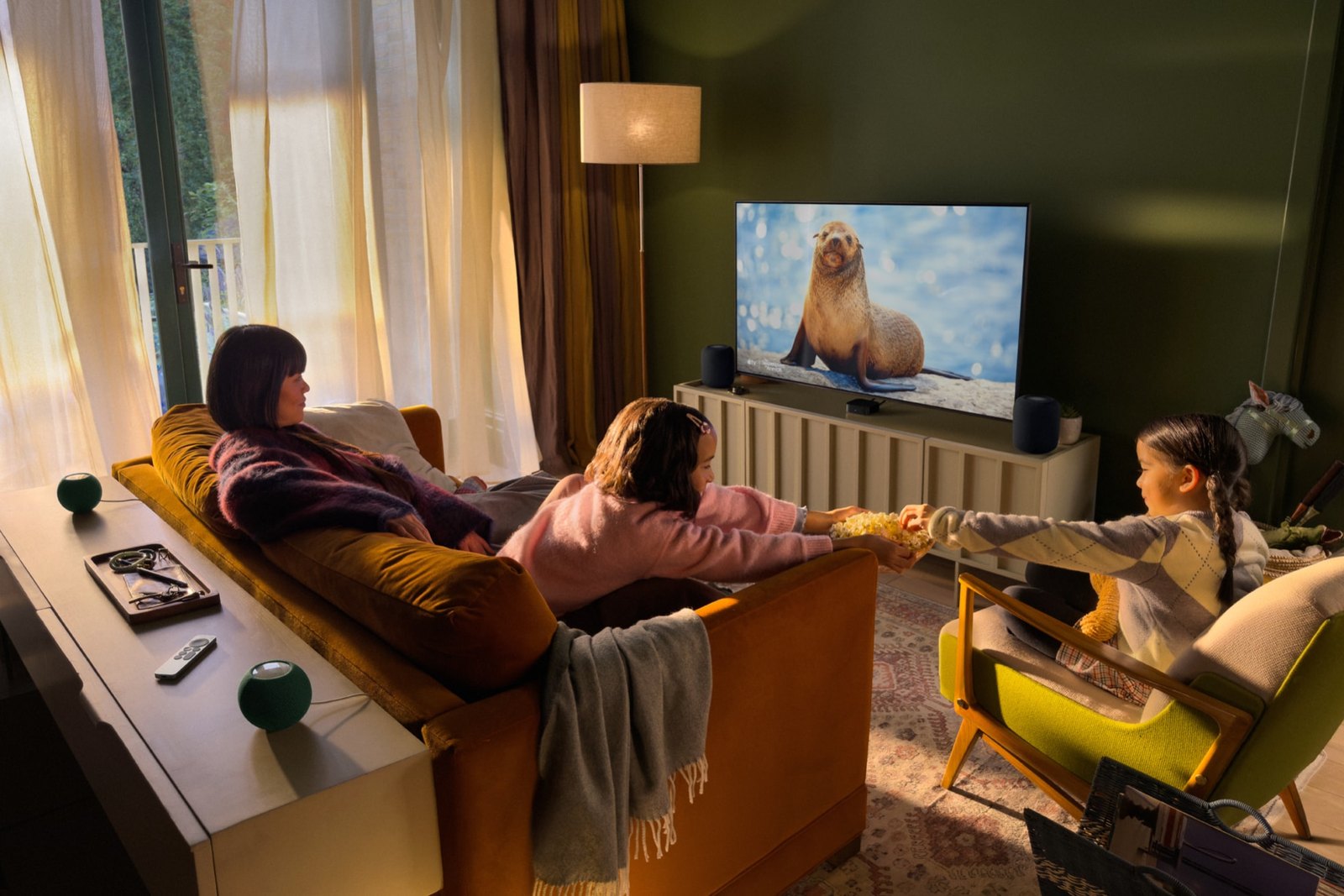 Famille dans un salon devant une télévision, avec l'Apple TV 4K sous l'écran, deux HomePod mini noirs près du téléviseur et deux verts près du canapé.